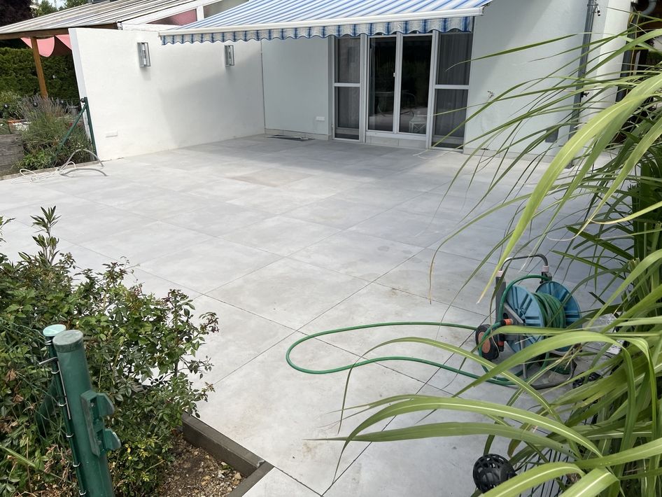 Große gepflasterte Terrasse vor einem weißen Haus mit Glas-Schiebetür und grünem Gartenschlauch auf dem Boden