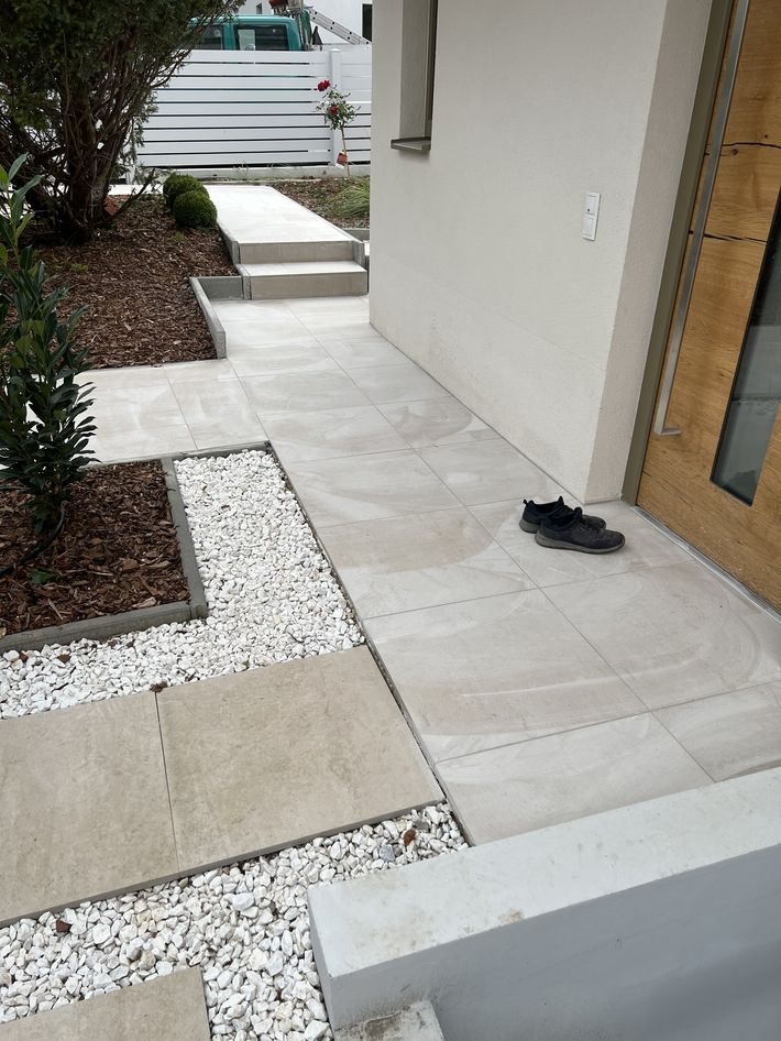 Eingangsbereich mit hellen Bodenplatten, weißen Ziersteinen, einem Paar schwarzer Schuhe neben der Haustür und einem Weg mit Stufen im Garten