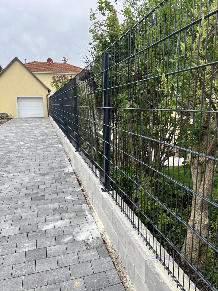 Drahtzaun auf einer niedrigen Mauer entlang eines gepflasterten Wegs mit Bäumen dahinter und einem Haus im Hintergrund