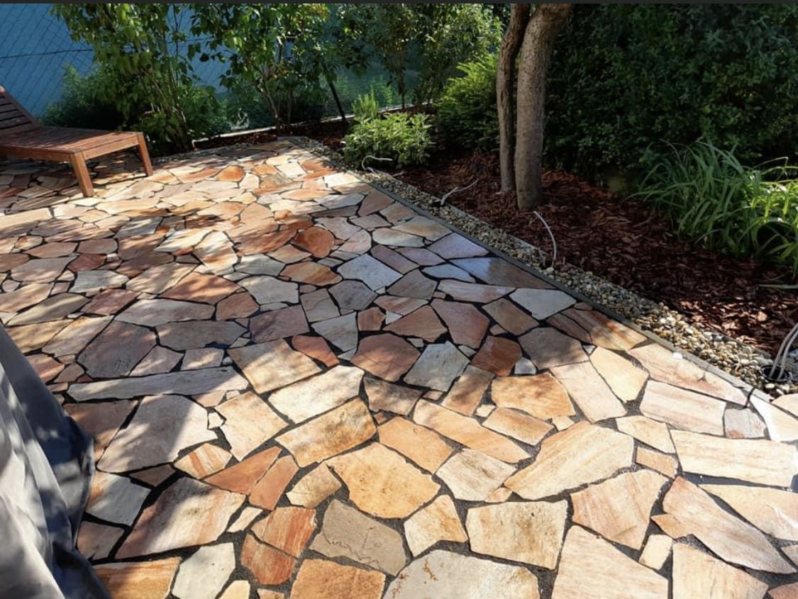 Gepflasterter Boden aus unregelmäßigen Natursteinplatten in einem Garten mit Bäumen und einer Holzbank im Hintergrund