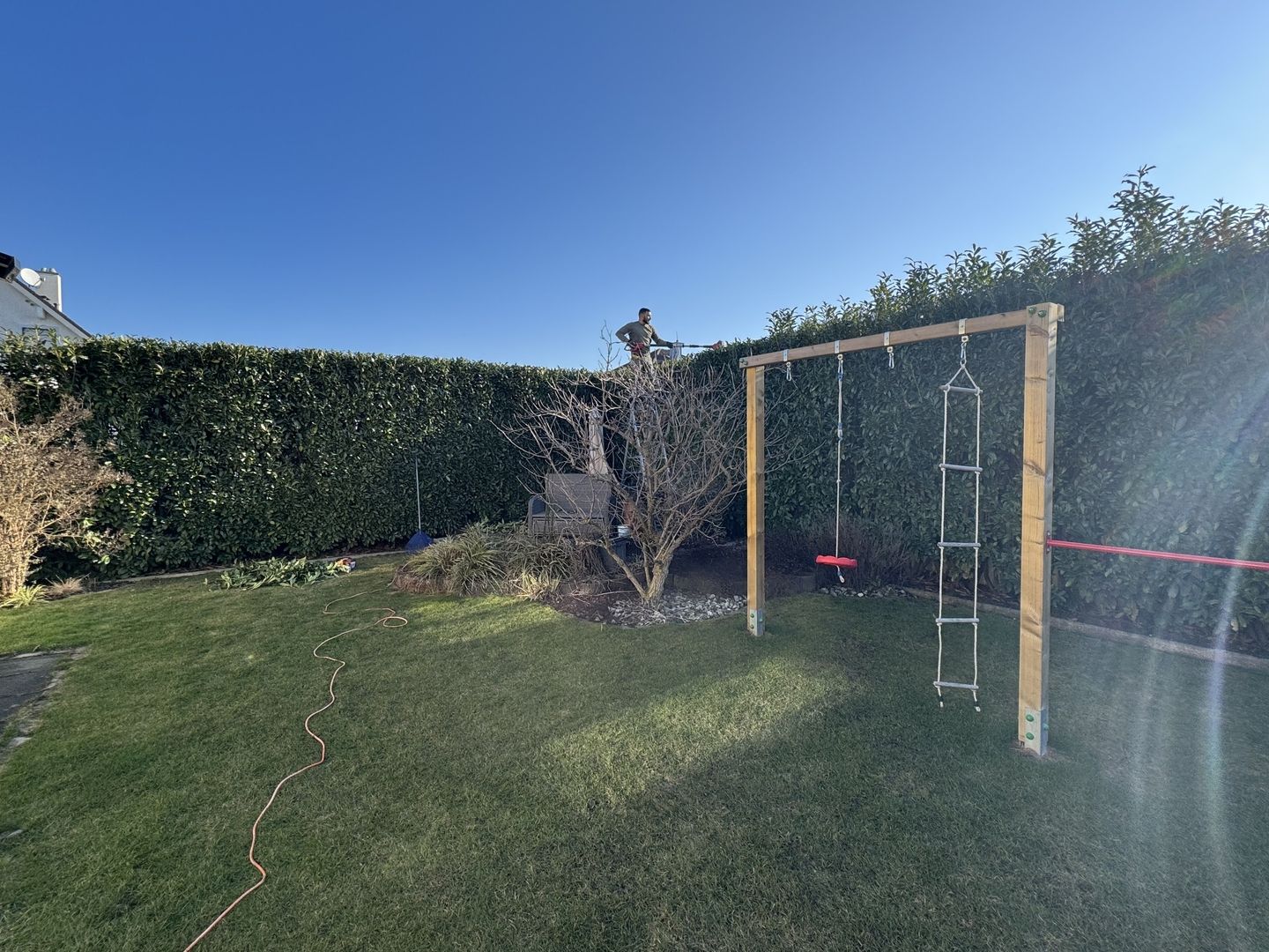 Garten mit grünem Rasen, Hecke im Hintergrund, einem Klettergerüst mit Leiter und Schaukel sowie einem Baum mit kahlen Ästen